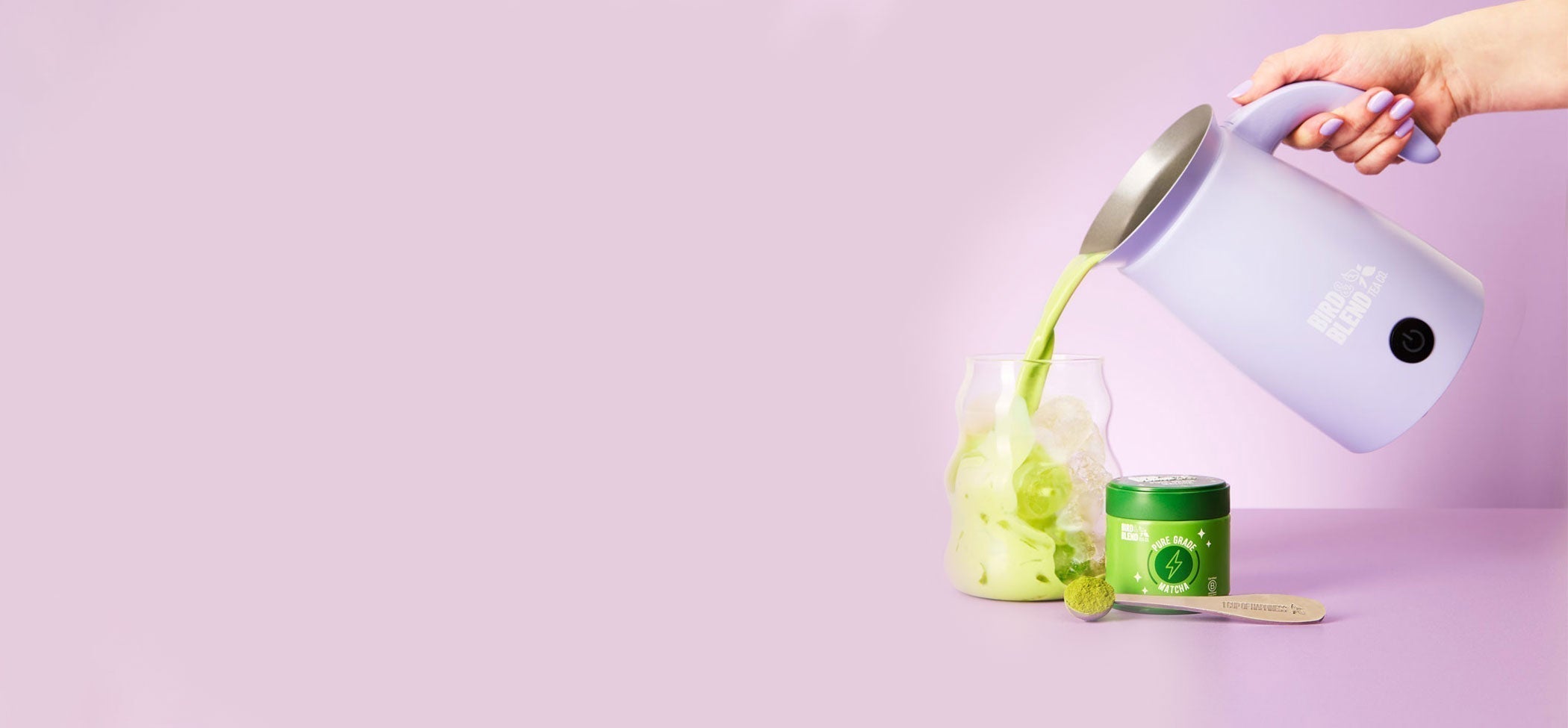 matcha pouring from a soft lilac matcha latte magic whisk in a glass with ice and pure grade matcha and spoon filled with matcha powder