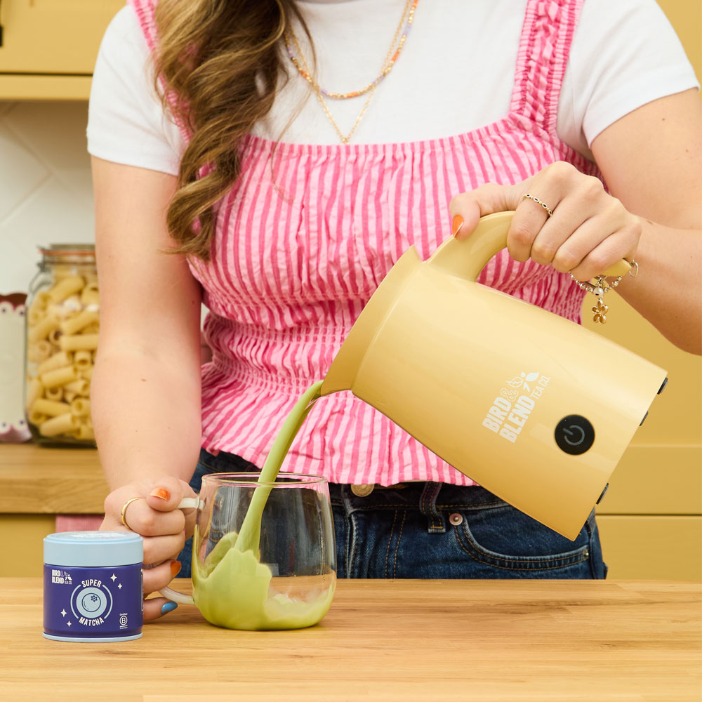 blueberry &amp; acai super matcha tin and person pouring hot blueberry &amp; acai super matcha from a bamboo gloss matcha latte magic whisk